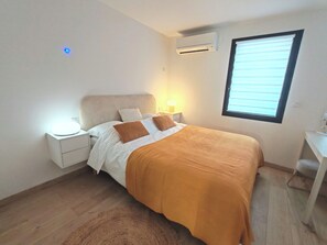 Ein Schlafzimmer mit Bett, Nachttisch, Lampe, Klimaanlage, Fenster und einem kleinen Schreibtisch.