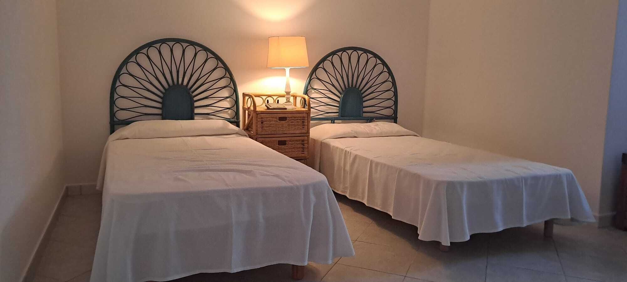A room with two single beds, each with a headboard and a bedside table with a lamp.