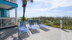 Une terrasse en bois avec des chaises longues bleues, un palmier et un panneau indiquant : « PROTEGEZ LA LIGNE DE DUNES. »