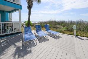 A wooden deck with blue lounge chairs, a palm tree, and a sign that reads 'KEEP THE DUNE LINE.'