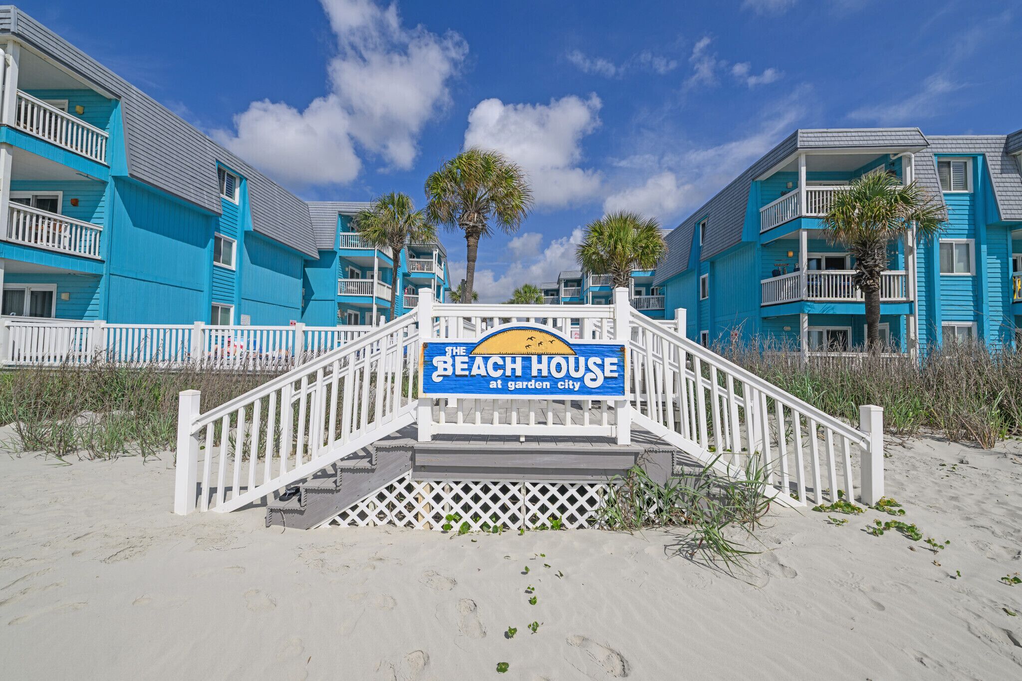 A beachfront hotel with a sign reading 'The Beach House at Garden City,' featuring a white staircase leading to the entrance, surrounded by palm trees and a clear sky.