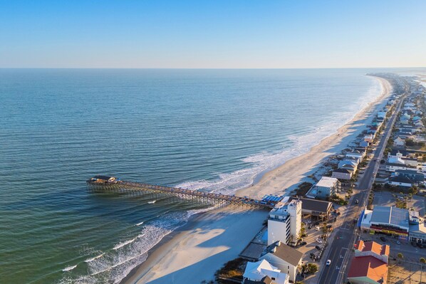 An aerial view of a coastal town with a pier extending into the sea, adjacent to a sandy beach and lined with buildings.