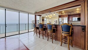 A bar area with a view of the sea, wooden chairs, and a kitchen with wooden cabinets. - 🌞Sunsets, Gulf and Beach Views🐬Great Amenities🌴Mansions By The Sea 1103A (Treasure Island)