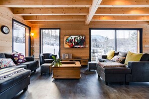 Ein gemütliches Wohnzimmer mit Holzwänden und großen Fenstern, ausgestattet mit einer Couch, Sesseln, einem Couchtisch und einem Fernseher.