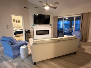 Ein Wohnzimmer mit Kamin, einem darüber montierten Fernseher, einem Deckenventilator und Blick durch eine Glastür ins Freie.