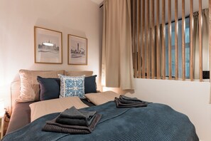 Ein ordentlich bezogenes Bett mit blauer Decke, zwei Kissen und einem Kissen mit blau-weißem Muster. An der Wand hängen zwei gerahmte Bilder, ein Fenster mit Holzjalousien und ein Nachttisch mit Lampe.