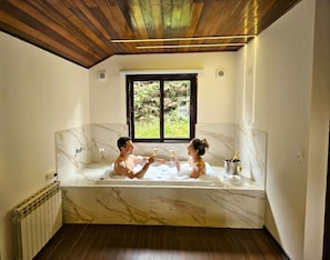A room with a large bathtub, a window with a view of greenery, and a radiator.
