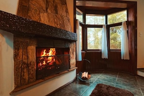 A stone fireplace with a fire, a window with curtains, and a corner rug. - Pousada Recanto dos Sonhos (Campos do Jordão)