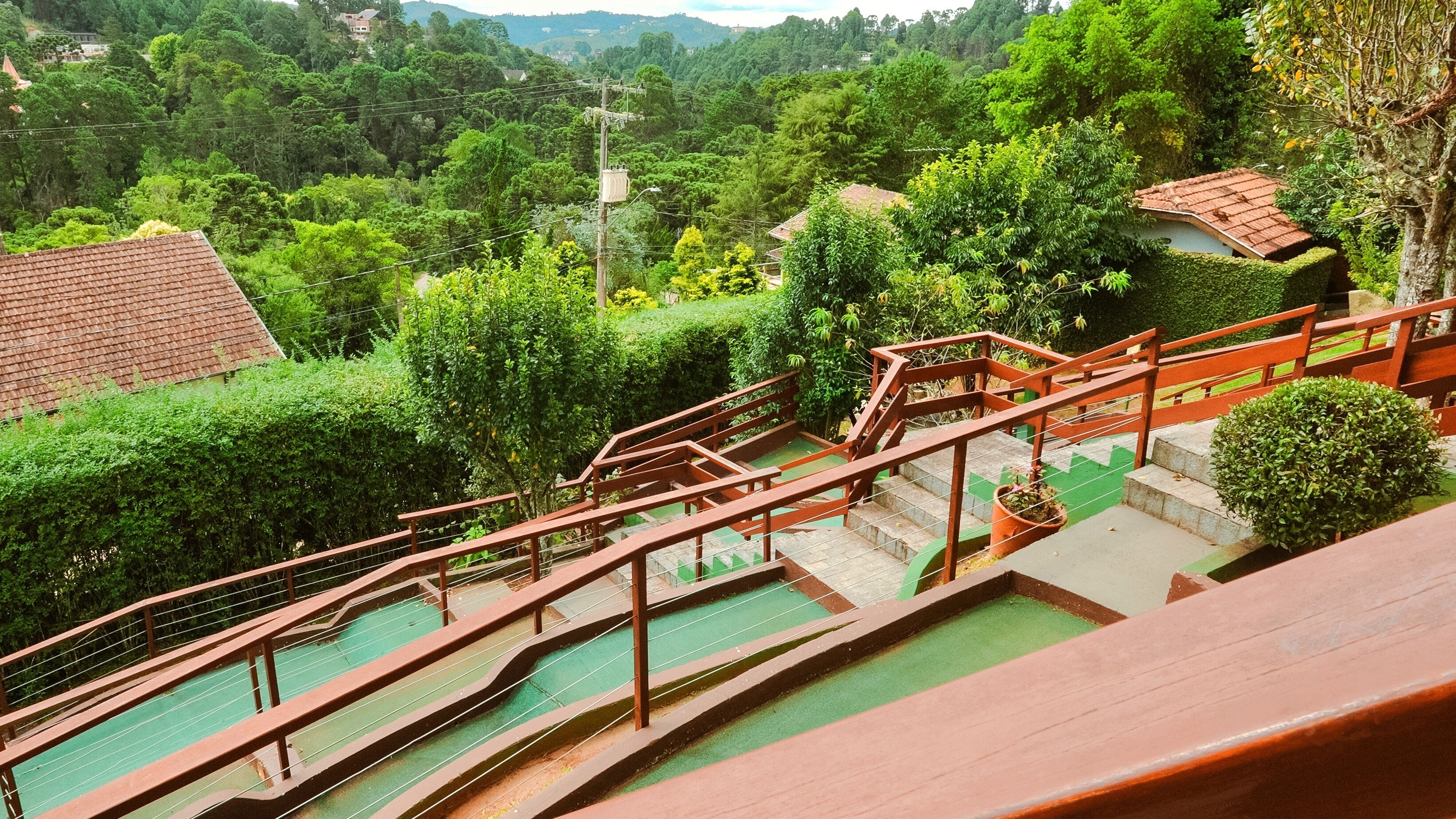 A staircase with a green safety surface leading to a higher level, surrounded by lush greenery and a view of a hilly landscape.