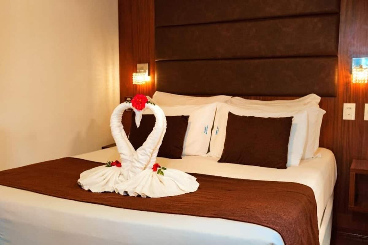 A hotel room with a neatly made bed, a decorative swan towel, and two wall-mounted lamps.