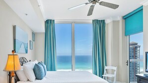 Une chambre d’hôtel avec un lit, un ventilateur de plafond, une vue sur l’océan et un balcon donnant sur la ville.