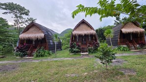 Tres cabañas con techo de paja y balcones de madera, rodeadas de vegetación exuberante y un sendero.
