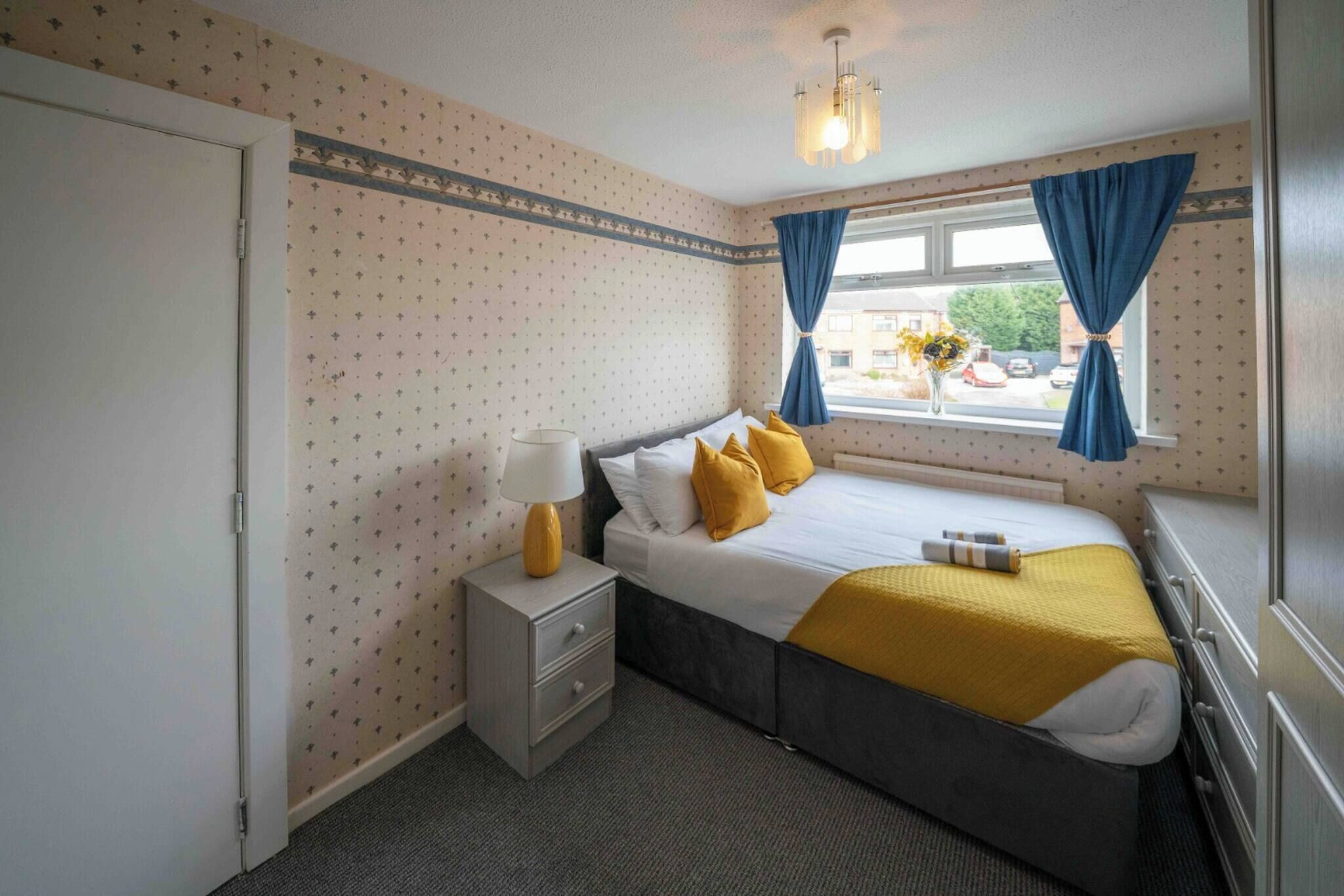 A bedroom with a bed, a nightstand, a window with curtains, and a view of the outside.