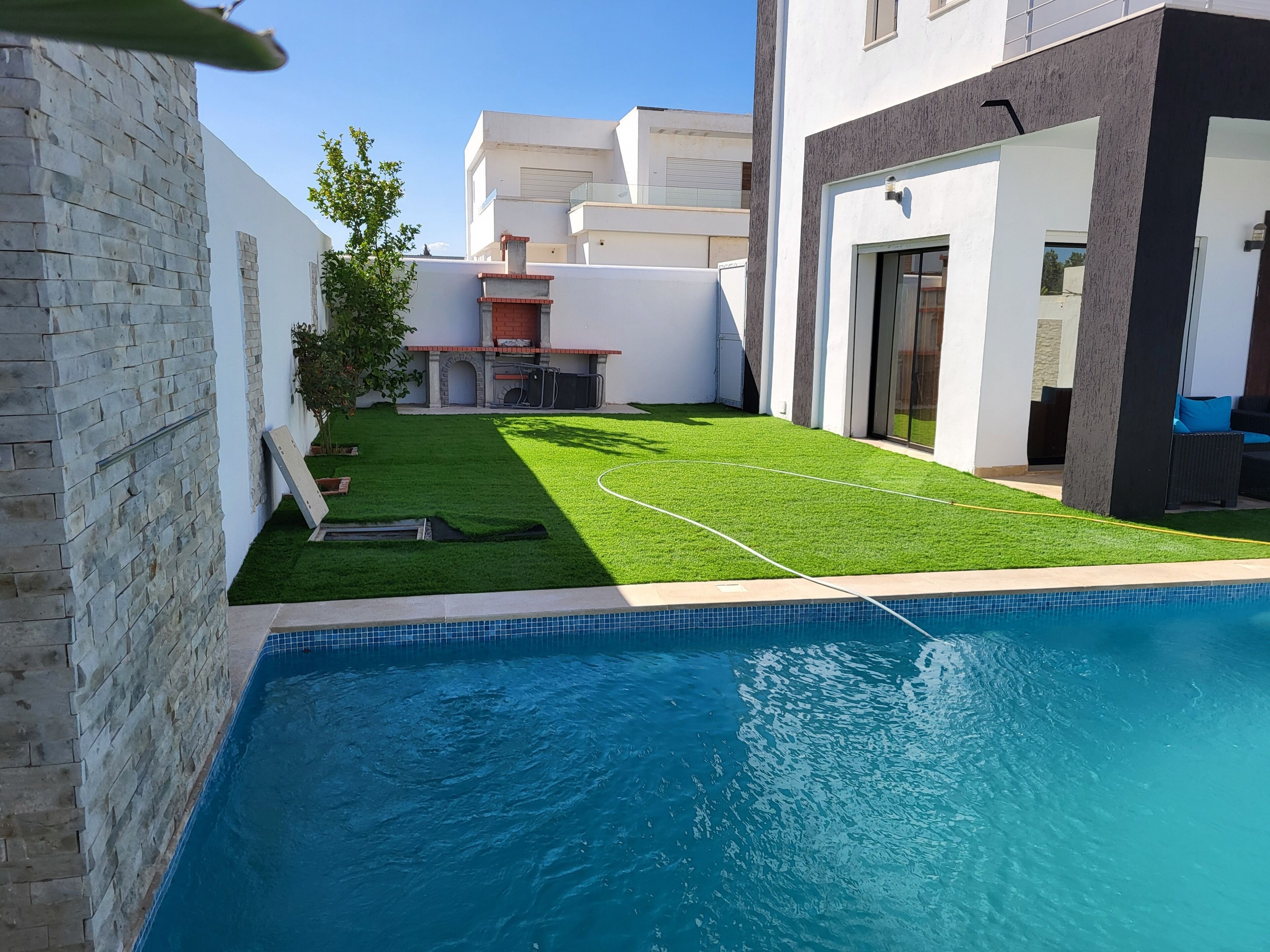 Une piscine avec un bord carrelé, une petite pelouse avec du gazon synthétique et un mur en briques.