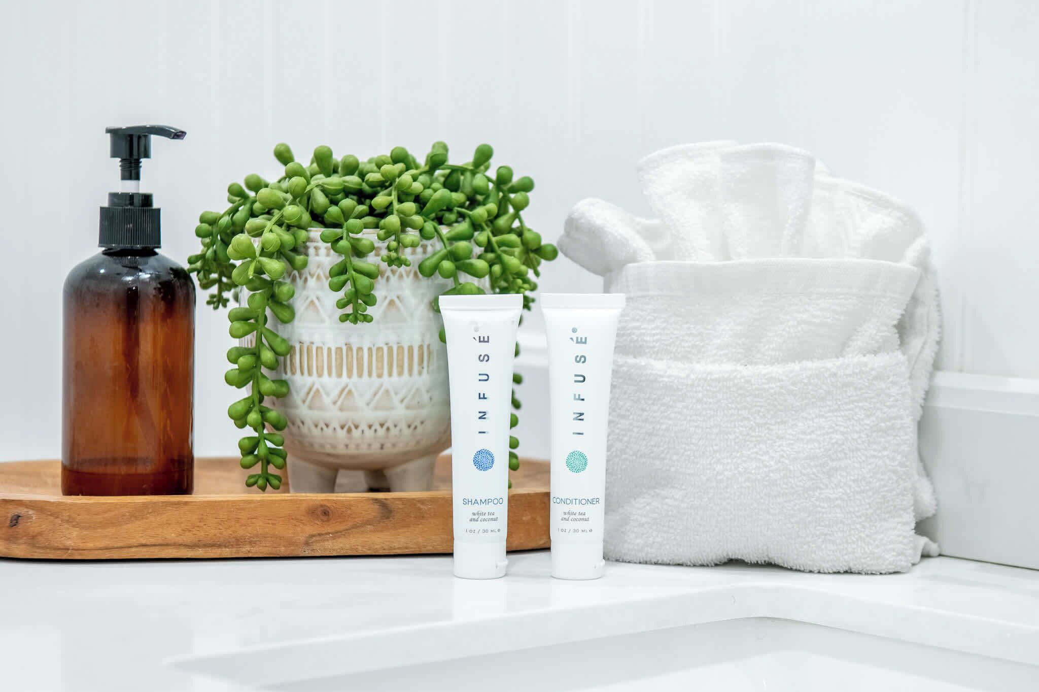 A bathroom with a wooden tray holding a brown bottle, two white tubes labeled 'INFUS,' a white towel, and a green plant.
