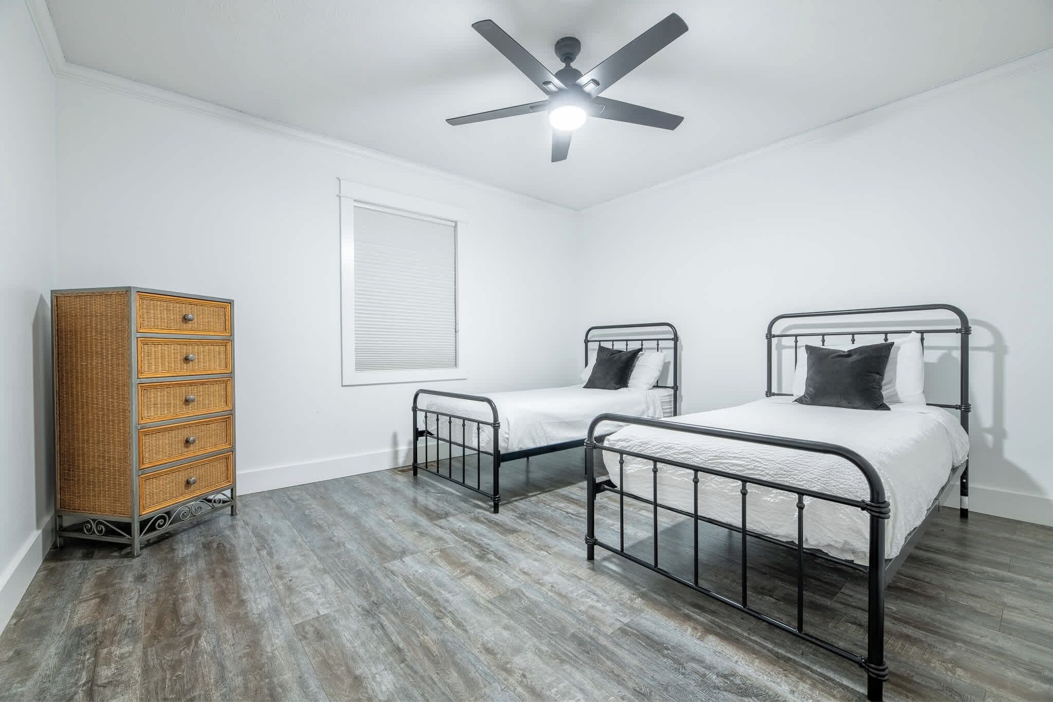 Quarto com duas camas de metal, cômoda de madeira, ventilador de teto e janela com persianas.
