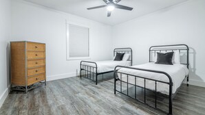 Un dormitorio con dos camas de metal, un aparador de madera, un ventilador de techo y una ventana con persianas.