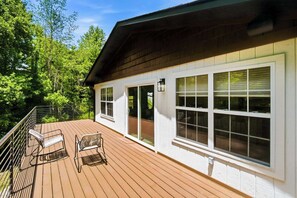 Eine Holzterrasse mit zwei Stühlen, ein Gebäude mit großen Fenstern und ein grünes, bewaldetes Gebiet.