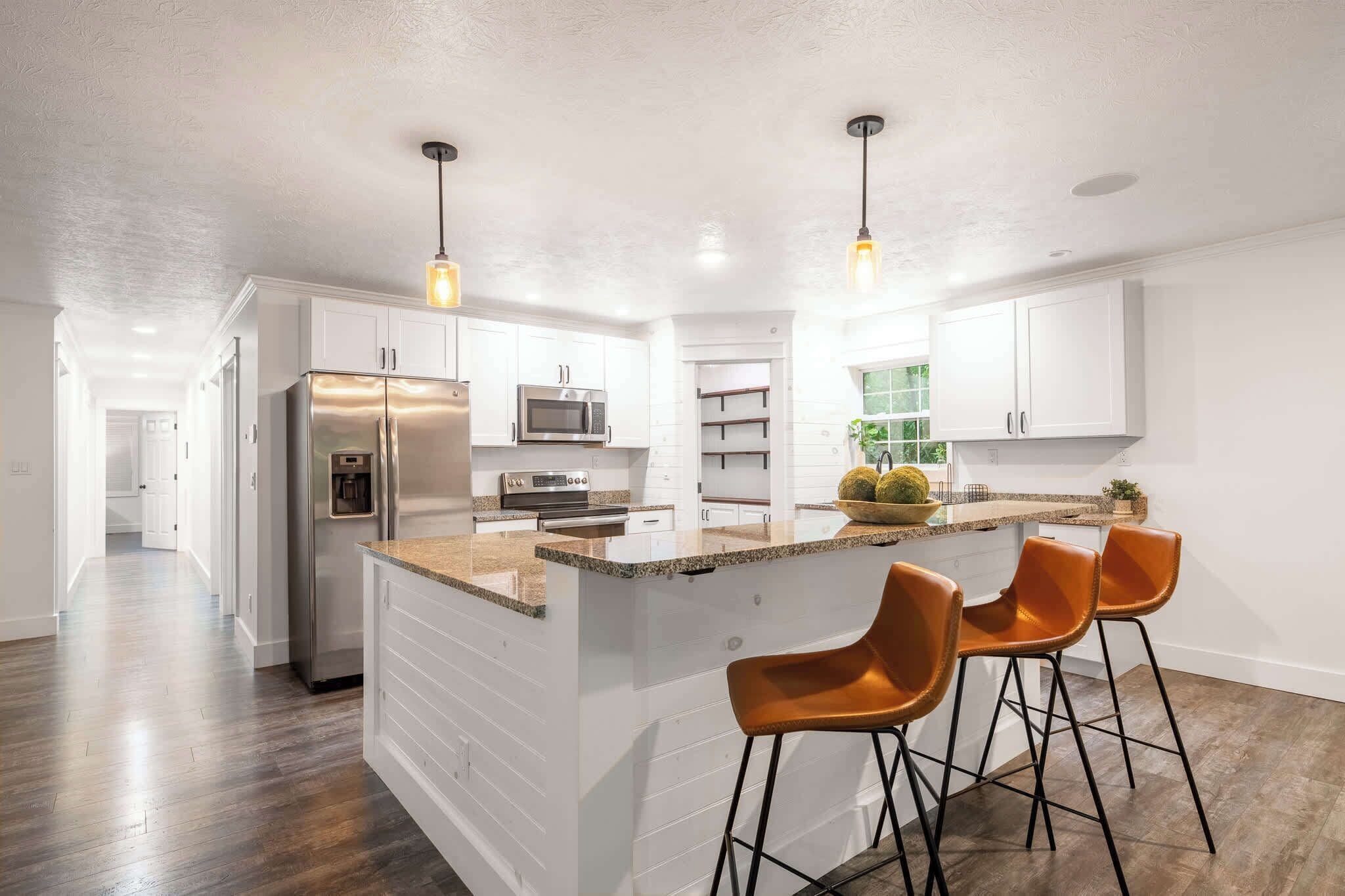 A modern kitchen with white cabinetry, a stainless steel refrigerator, a microwave, and a built-in oven. There are two bar stools with black legs and a wooden island with a granite countertop.