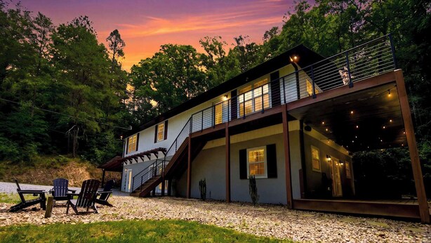 Una casa de dos pisos con un área de estar cubierta al aire libre, una escalera y un amplio ventanal con vistas a un bosque.