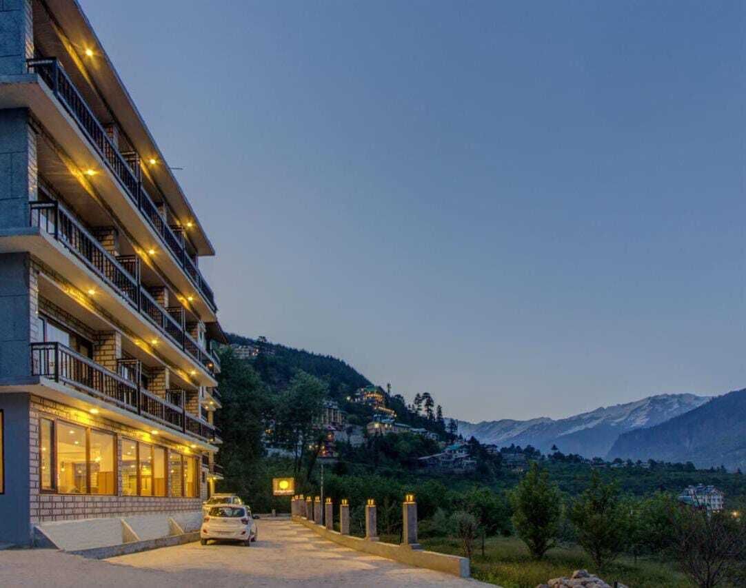 A multi-story hotel with balconies, a car parked in front, and a mountainous backdrop.