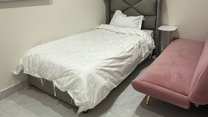 Una cama individual con cabecera tapizada de gris, una mesita de noche y una silla rosa en una habitación con suelo de baldosas.