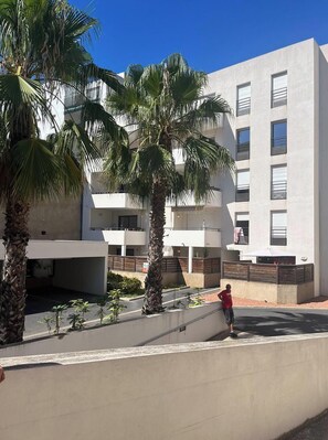 A modern building with multiple balconies, palm trees, and a paved area with a person standing.