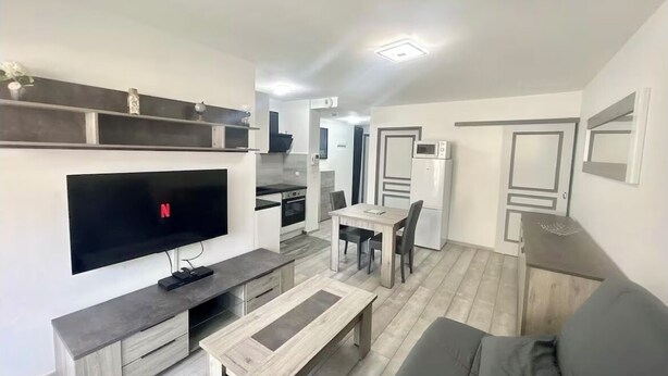 A modern living room with a flat-screen TV, a wooden coffee table, a gray sofa, and a dining area with a table and chairs. - Studio parking clim wifi parking downtown (Béziers)