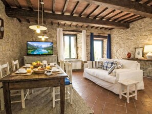 Ein rustikaler Raum mit Steinwand, Holzbalkendecke, einem Esstisch mit Frühstück, einem weißen Sofa und einem Fernseher, der eine Berglandschaft zeigt.