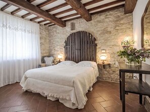 Ein Schlafzimmer mit Steinwand, Holzbalken, einem Bett mit weißer Bettwäsche, zwei Nachttischlampen und einem kleinen Tisch mit Blumenvase.