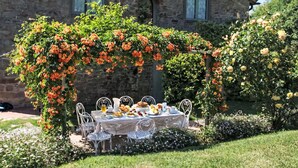 Ein Steinhäuschen mit Garten und Terrassensitzplatz, an dem ein Tisch für eine Mahlzeit gedeckt ist, umgeben von blühenden Pflanzen.
