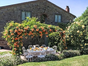 Ein Steinhäuschen mit Garten und Terrassensitzplatz, an dem ein Tisch für eine Mahlzeit gedeckt ist, umgeben von blühenden Pflanzen.