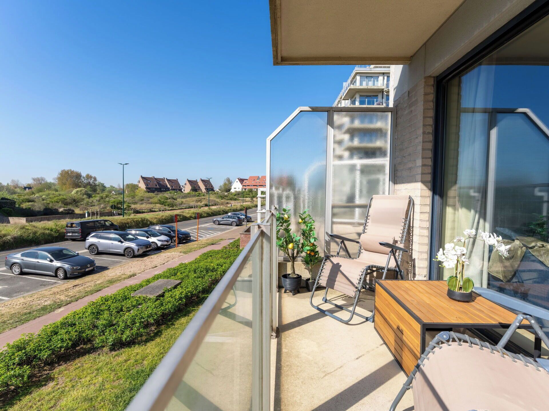 En balkon med udsigt til en parkeringsplads, et træbord, en stol og en potteplante.
