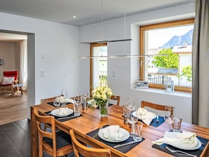 Un coin repas avec une table en bois dressée pour quatre personnes, un vase de fleurs et une vue sur les montagnes par la fenêtre.