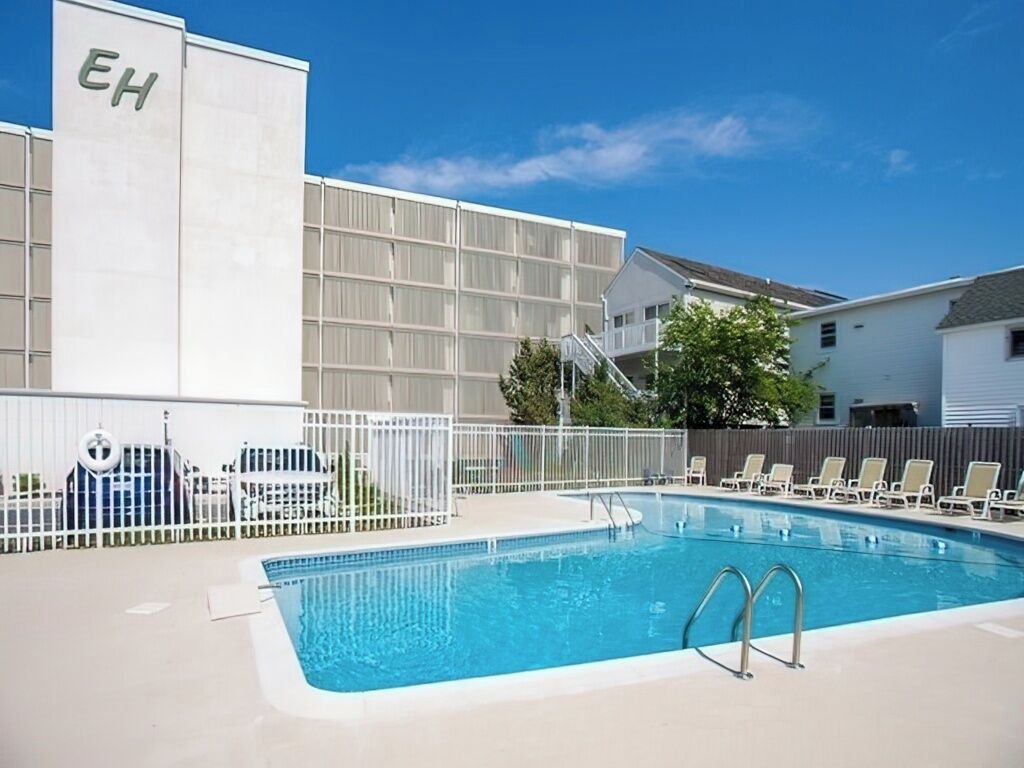 A swimming pool with a white fence, lounge chairs, and a building with 'EH' on its side.