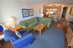 Ein Wohnzimmer mit grünem Sofa, blauem Sessel, Holztisch und einem Essbereich mit Tisch und Stühlen.