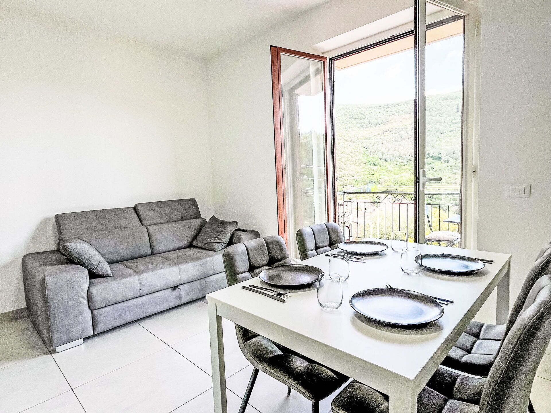 Un área de comedor con una mesa dispuesta para cuatro personas, un sofá gris y un ventanal con vistas a zonas verdes.