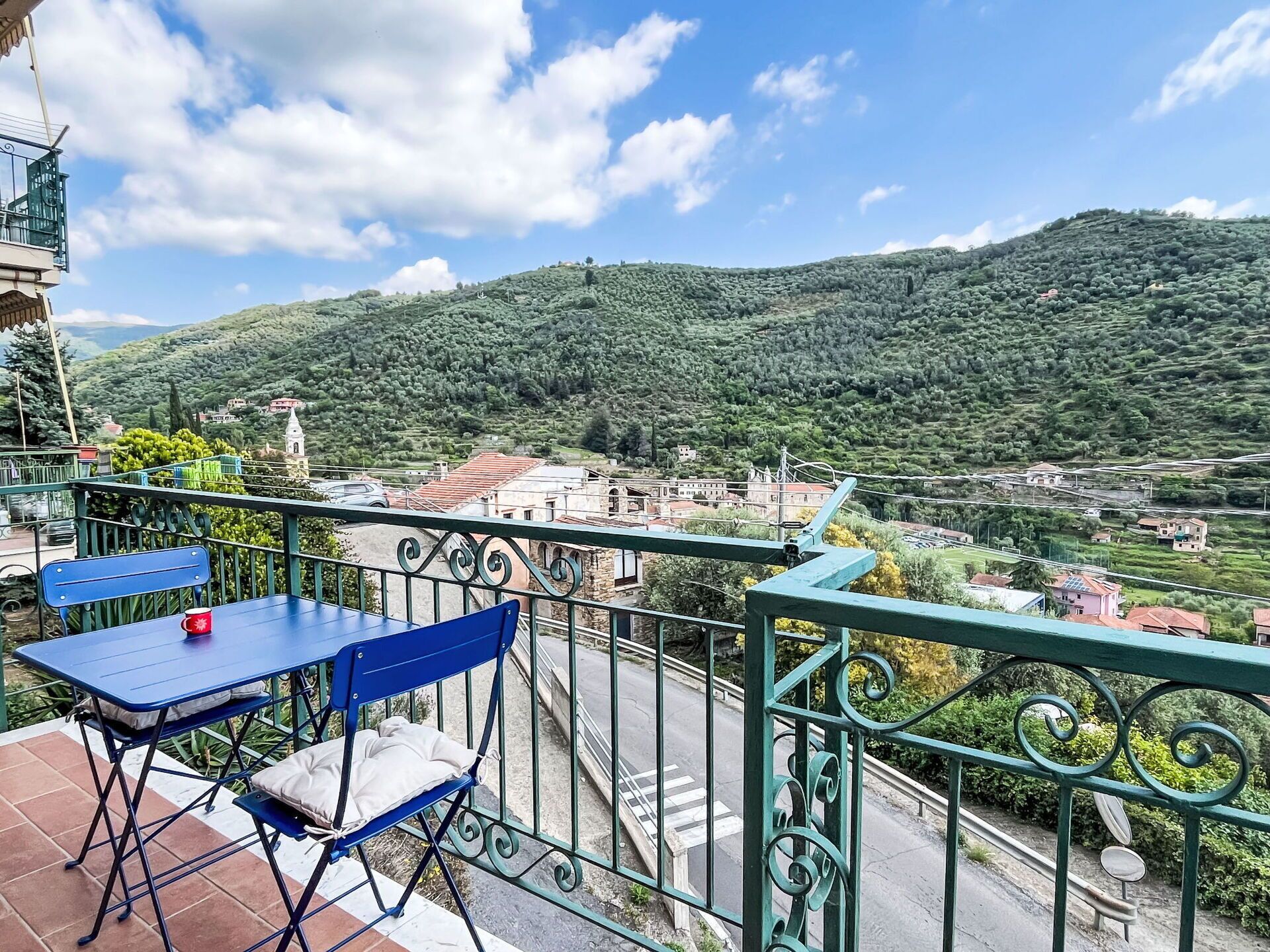 Ein Balkon mit Tisch und Stuhl, mit Blick auf ein Dorf und grüne Hügel.