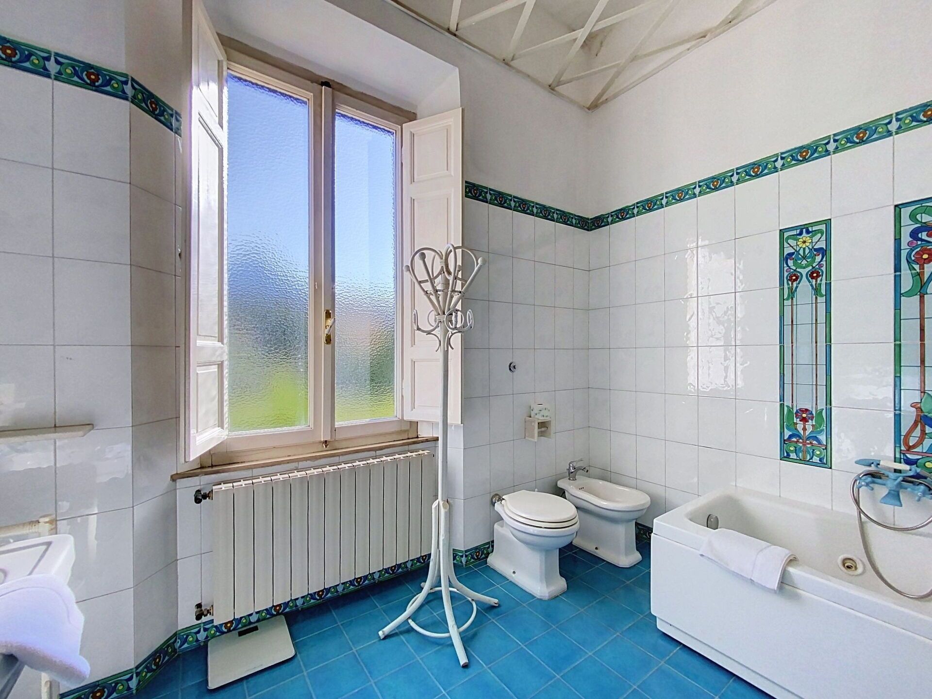 A bathroom with a window, two toilets, a bathtub, and a radiator.