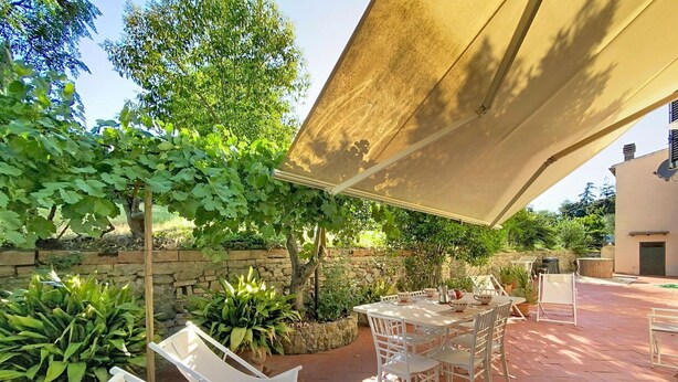 Un patio al aire libre con mesa y sillas, rodeado de follaje y un muro de piedra.