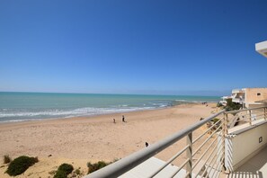 Blick auf einen Strand von einem Balkon mit Geländer, klarer Himmel und ruhiges Meer.