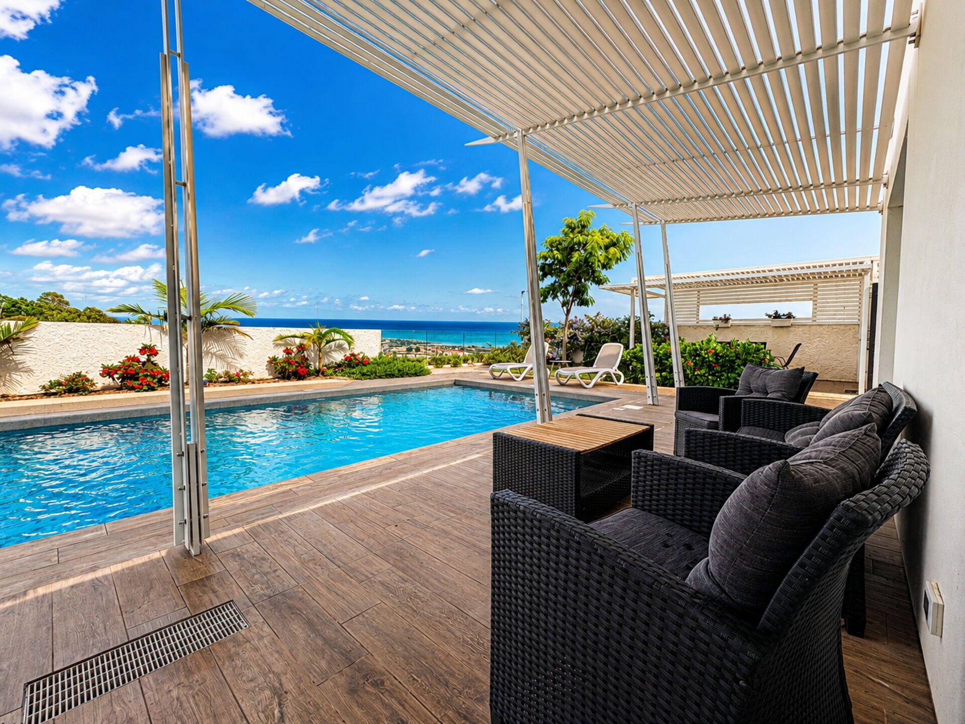 Zona junto a la piscina con vista al océano, mobiliario exterior y una pérgola.