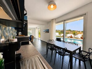A modern kitchen with a large island, a dining area with a table and chairs, and a view of a swimming pool. - Stunning private villa for 7 people with WIFI, A/C, private pool, patio and panoramic view (Marina Di Ragusa)