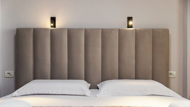 Ein Bett mit gepolstertem Kopfteil, zwei Nachttischlampen und einer neutralen Tagesdecke.