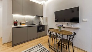 Eine kompakte Küche mit integriertem Mikrowellenherd, einer Kaffeemaschine und einem Spülbecken. Es gibt eine kleine Essecke mit einem Tisch und zwei Stühlen. Ein Flachbildfernseher ist über der Essecke an der Wand montiert.