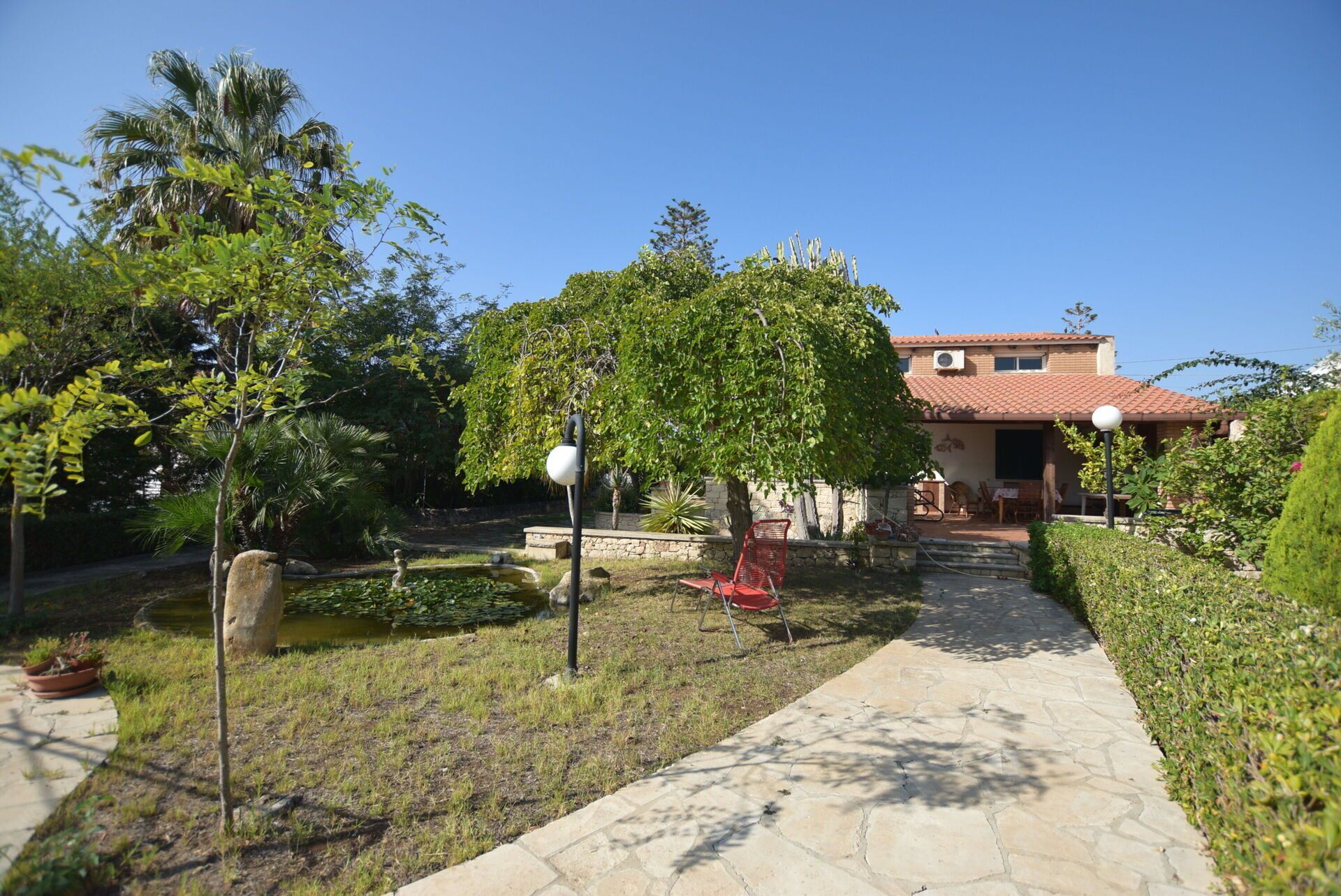 A garden pathway leading to a house with a tiled roof, surrounded by lush greenery and a small pond.