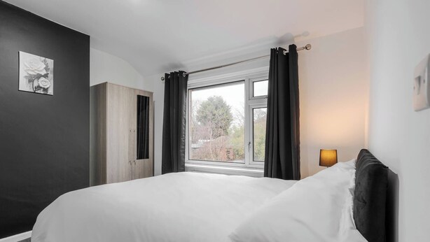 Un dormitorio con cama, ventana con cortinas, lámpara de noche y un cuadro en la pared.