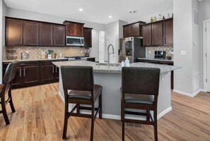 A modern kitchen with dark wood cabinets, a granite countertop, stainless steel appliances, and two bar stools. - 3BR Spacious Home with BBQ Patio,Backyard,Sleeps 6 (Aurora)