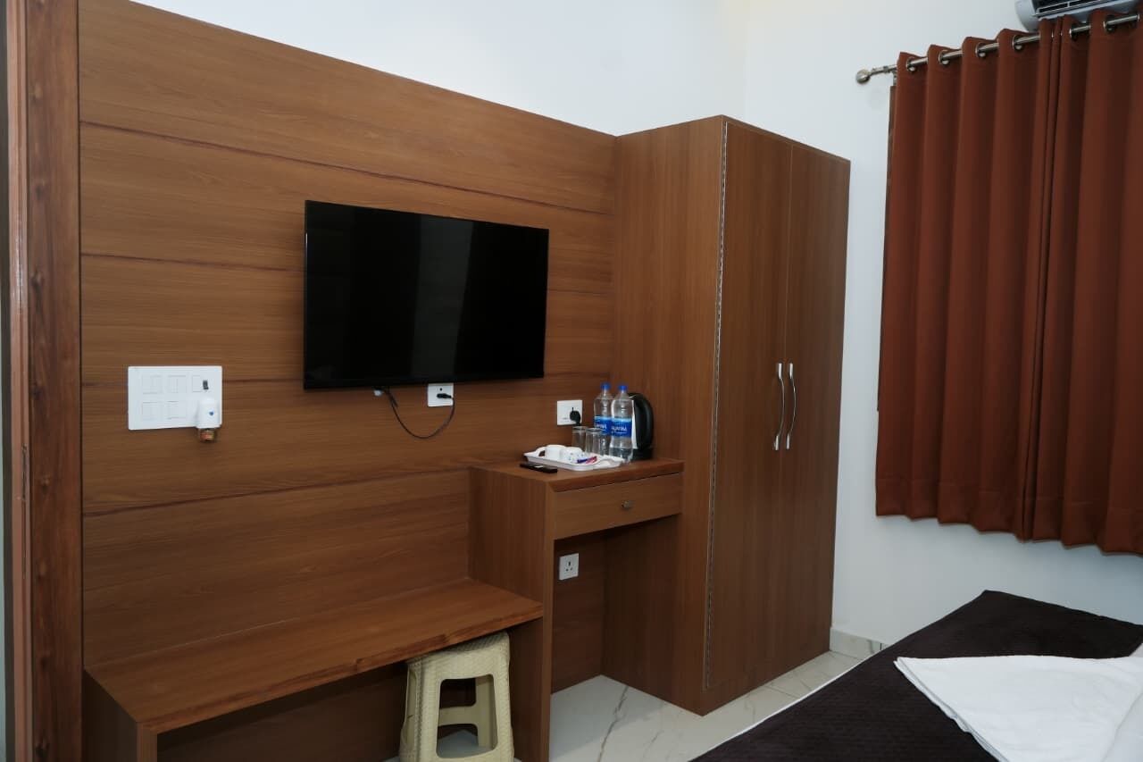 A hotel room with a wooden wardrobe, a desk with a flat-screen TV, a small stool, and a bed with a black and white blanket.