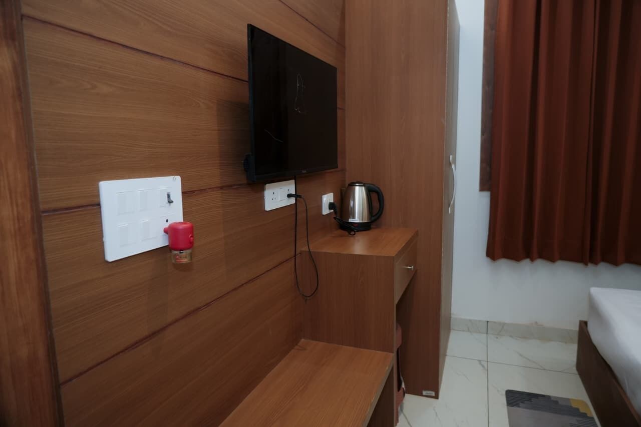 A hotel room with a wooden wall panel, a flat-screen TV mounted above a power outlet, a kettle on a wooden side table, a red cup on the wall, and a bed with a white mattress.